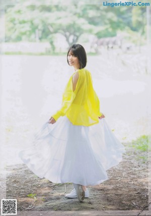 A woman in a yellow top and white skirt standing in the woods.