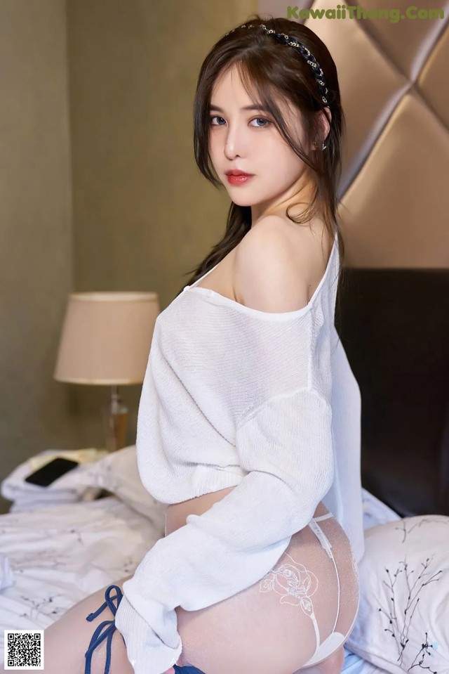 A woman in a white sweater and blue panties sitting on a bed.