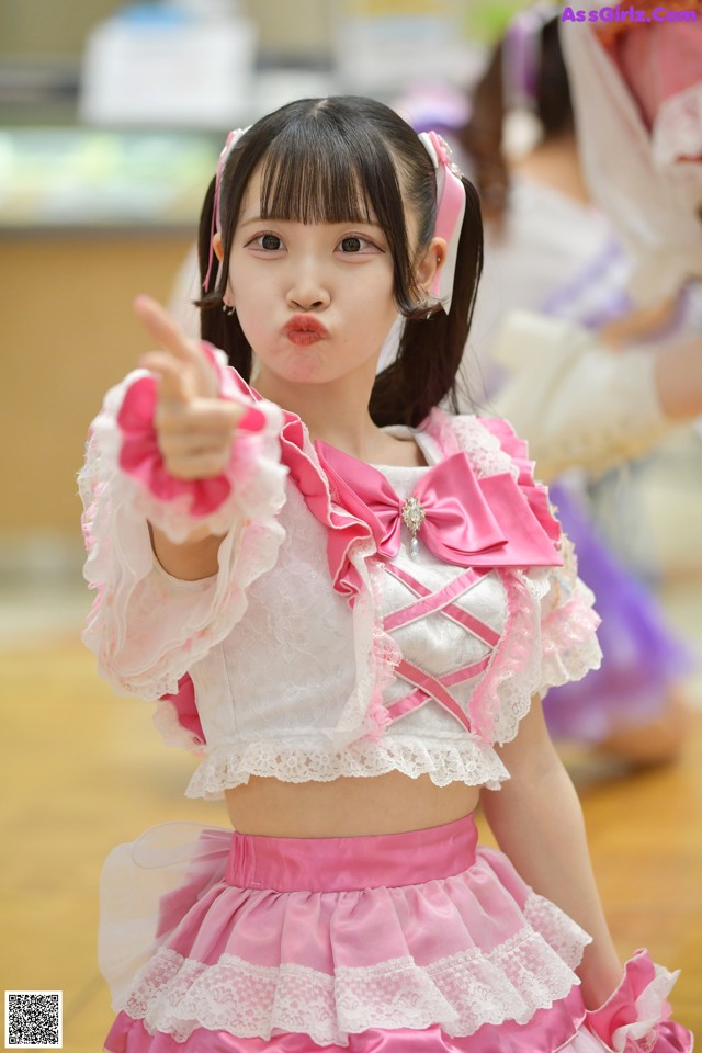 A young girl in a pink and white outfit pointing at the camera.