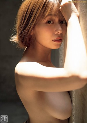 A naked asian woman leaning against a wall with her hands behind her head.